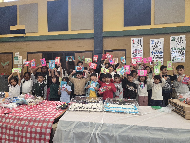 A diverse group of elementary school students proudly hold up colorful handmade cards during a classroom celebration or community event in a school cafeteria. Tables in front of the children display sheet cakes, pizza boxes, and supplies, creating a festive atmosphere focused on creativity, friendship, and student engagement.