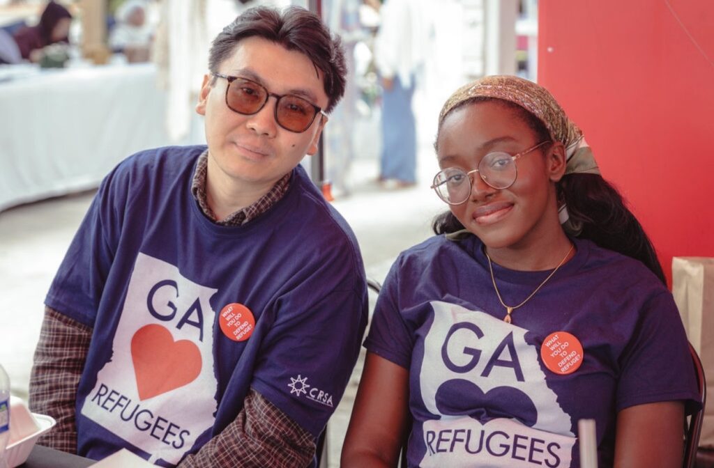 Two community members wearing matching "GA Loves Refugees" T-shirts sit together at a community event, smiling toward the camera. Both wear red buttons promoting refugee support, representing advocacy and solidarity with refugee communities in Georgia.
